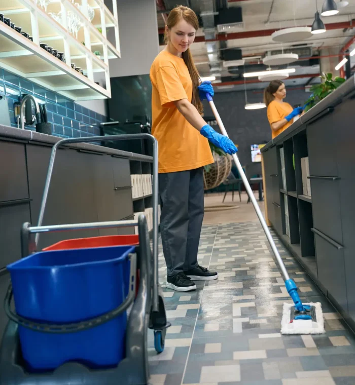 Young woman cleaning the floor with a mop, her colleague is in the background